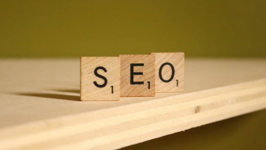 Three wooden Scrabble tiles spell out "SEO" on a wooden surface against a green background.