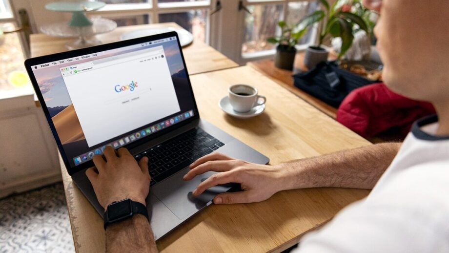 A person using a laptop with the Google search page open on the screen. There is a cup of coffee, plants, and other items on the wooden table in a well-lit room.