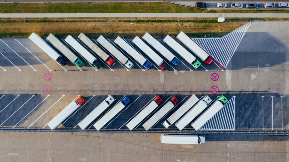 Aerial view of parked semi-trucks in angled parking spaces at a lot. A road with a few cars is visible at the top of the image.