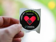 Hand holding a sticker that reads, I got my COVID-19 vaccine StopCOVID with a heart design and matrix logo.